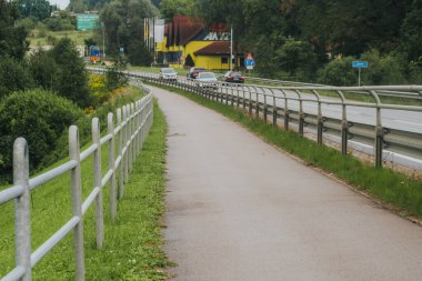 Metal korkuluklar ve yoğun yeşil ormanlarla çevrili kaldırımlı bir yolda, paralel yaya yolu ve yol kenarı işaretleri olan bir araç sürüyor..