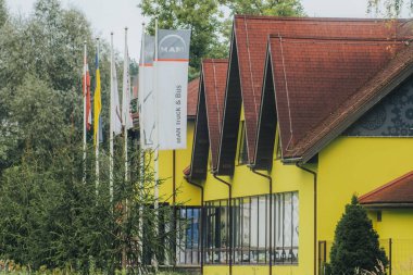 Üçgen kapaklı çatılı canlı sarı bir binada yosunlu fayanslar ve birkaç damgalı bayrakla çevrili büyük pencereler bulunur..