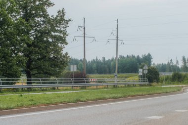 Metal bariyerlerle çevrili kıvrımlı bir kırsal yol, elektrik direkleriyle ve tepedeki elektrik hatlarıyla yeşil bir arazi boyunca uzanıyor. Sahne yön tabelaları ve ormanlık bir arka plan içeriyor, sakin bir kırsal ortam yaratıyor.