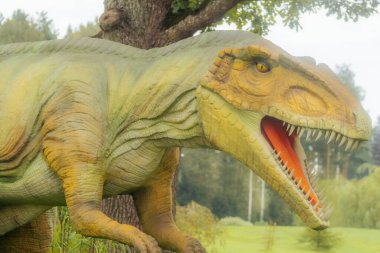 Açık ağızlı ve keskin dişli yeşil ve sarı dinozor heykeli yemyeşil bir parkta duruyor..