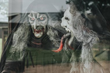 İki ürkütücü cadı figürü, soluk tenli, ağzı kanlı, keskin dişli ve uzun saçlı korku temalı bir ekranda camın arkasında duruyor.. 