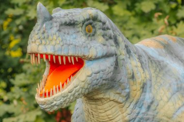 Keskin dişli ve turuncu ağızlı mavi ve sarı bir dinozor heykelinin yeşil yapraklara karşı detaylı görüntüsü.. 