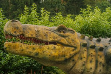 Ağzı açık ve keskin dişleri olan detaylı bir dinozor heykeli yoğun yeşil bitkiler arasında duruyor, dokulu derisi ve canlı görünümlü tarih öncesi bir görünüşü var..
