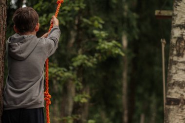 Gri kapüşonlu bir çocuk ormanlık bir alanda parlak turuncu bir ipe tutunarak uzun ağaçların arasında bir etkinliğe hazırlanıyor..
