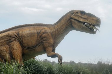 Keskin dişli büyük gerçekçi bir Tyrannosaurus Rex heykeli, bulutlu bir gökyüzü arka planına karşı çenesinde başka bir dinozor modeli tutuyor.. 