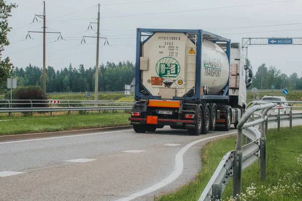 Silindirik bir tankı olan bir gaz taşıyıcısı Mujani 'yi işaret eden bir yol tabelasının yakınındaki dolambaçlı bir yolda ilerliyor. Ağaçlarla, çimenli bölgelerle ve elektrik direkleriyle kırsal alan.