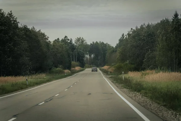 Bir araba, bulutlu bir gökyüzünün altında çimenli tarlalar ve sık ormanlarla çevrili sakin iki şeritli bir kır yolu boyunca ilerliyor.. 