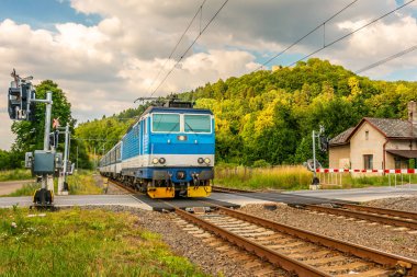 Geçiş engelleri ile gelen mavi elektrikli tren. Yeşil vadi boyunca çalışan bir tren. Taşıma Çek Cumhuriyeti'nde demiryolu. Demiryolu güneşli bir günde