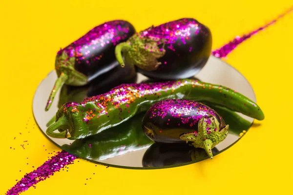 Composition of eggplants and peppers on round mirror covered by glitter.