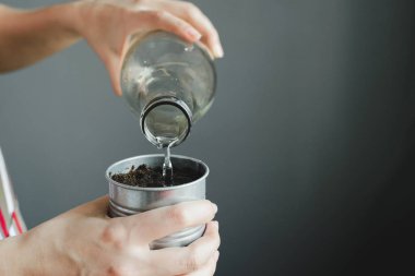 Kadınların elleri cam şişeyi tutuyor ve toprağı metal tencerede suluyor. 