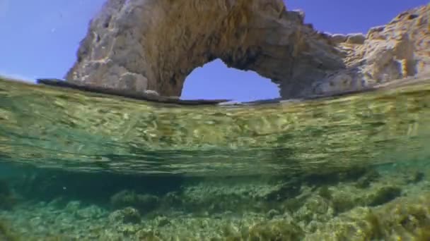 Camera steekt het wateroppervlak over, toont tegelijkertijd een rotsachtige bodem en kustrots in de vorm van een boog, gespleten. — Stockvideo