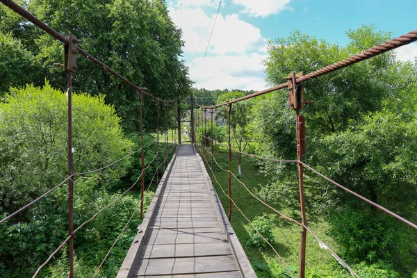 Tahta ve metal kablolardan yapılmış doğaçlama bir asma köprü sık ağaçların arasından geçiyor. Yaz güneşi yolu aydınlatırken, gökyüzü aydınlık bulutlarla dolu..