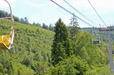 Yeşil ormanla kaplı bir dağ yamacının yaz panoramaları. Boş asansör koltukları temiz gökyüzüne karşı görünür, sizi yürüyüşe davet ediyor..
