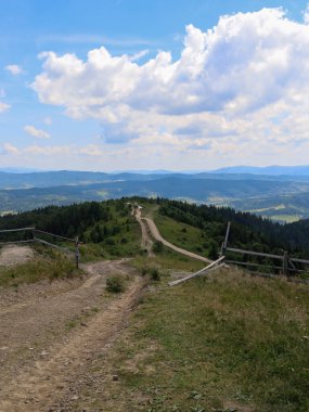 Dağ yamacı boyunca uzanan toprak bir yol sizi yeşil tepeler ve ormanlar arasında bir yolculuğa davet ediyor. Ufuktaki görkemli dağ sırtı ve uçsuz bucaksız bulutlu gökyüzü özgürlük ve huzur hissi veriyor..