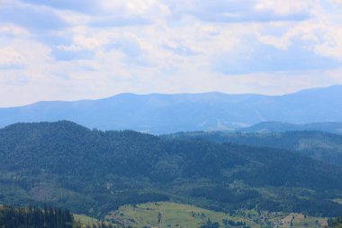 Ormanlı yamaçların mavi tepelere yol verdiği katmanlı bir dağ manzarası. Bu görüntü, uçsuz bucaksız mavi gökyüzü altında tüylü bulutlarla, doğanın sükunetini ve ihtişamını gösteriyor..