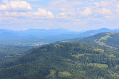 Ufuk çizgisine uzanan görkemli dağ sırtları manzarası katmanlı bir manzara yaratıyor. Yaz ormanları dağ yamaçlarını kaplıyor ve beyaz bulutlu mavi gökyüzü bu manzarayı huzur ve cazibe veriyor..
