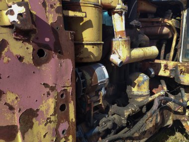 Eski bir traktörün paslı, bakımsız mekanik detaylarının makro fotoğrafı. Soyulmuş boya, pas izleri ve toprak zamanla çürümeyi simgeler. Güneş ışığı ise şekillerin ve dokuların karmaşıklığını vurgular., 