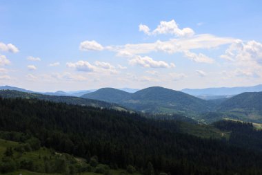 Sıcak bir yaz günü, sık ormanlarla kaplı görkemli dağların panoramik manzarasını ortaya çıkarıyor. Ufukta dağ zirveleri, tüylü beyaz bulutların tembelce sürüklendiği uçsuz bucaksız mavi gökyüzü ile kademeli olarak birleşir..