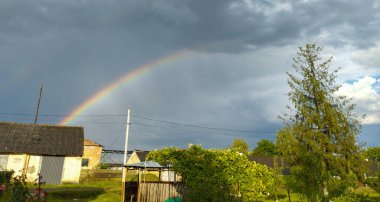 Yağmurdan sonra gökyüzünde parlak bir gökkuşağı olan pitoresk bir kırsal bölge. Kara bulutlar geri çekiliyor, güneş ışınları eski kır evlerini aydınlatıyor..