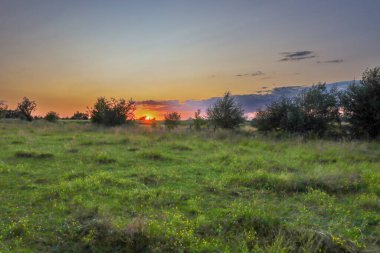 Açık bir arazide günbatımı olan sakin bir manzara, ufku turuncu ve pembe renklerle aydınlatıyor. Fotoğraf, sakin bir atmosfer ve günün sonunda kırsal kesimde güzel bir manzara yaratıyor..