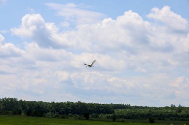 Yeşil tarlalardan ve orman kemerlerinden oluşan engin bir manzara geniş, mavi bir gökyüzü altında uzanıyor. Kabarık beyaz bulutlar altında yalnız bir kuş süzülüyor.. 