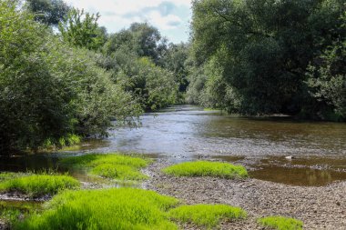 Bir nehir yavaşça kıyılarının yanından akar. Parlak yeşil çimenlerin toprak ve taşlardan oluşan bir zeminde yetiştiği yerden. Ağaçların ve çalıların çevreleyen yeşilliği güneş ışığıyla tamamlanmış sakin bir doğal atmosfer yaratır..