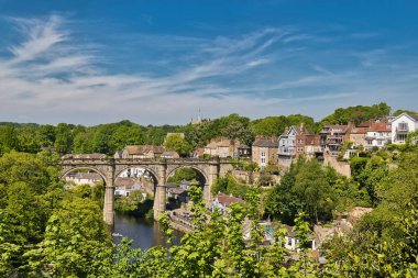 Resimli bir sahnede, Knaresborough, İngiltere 'deki açık mavi bir gökyüzünün altında nehir üzerinde evler ve bereketli ağaçlar bulunan bir taş viyadük sergileniyor..