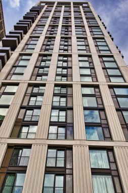 Bej cepheli ve koyu çerçeveli pencereli modern, uzun bir binanın alçak açılı görüntüsü. Balkonlar üst katlardan görülebilir. Londra, İngiltere 'de gökyüzü bulutlu..