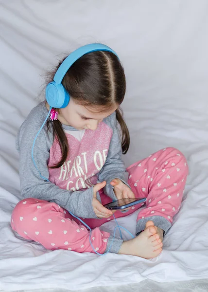 Mała Dziewczynka Niebieski Słuchawki Trzymając Rękach Inteligentny Telefon Siedzi Łóżku — Zdjęcie stockowe