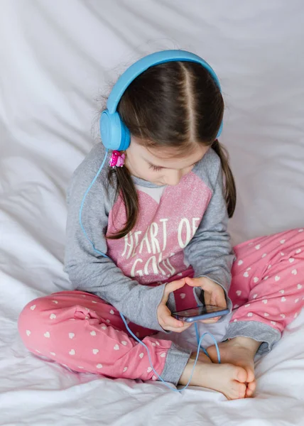 Mała Dziewczynka Niebieski Słuchawki Trzymając Rękach Inteligentny Telefon Siedzi Łóżku — Zdjęcie stockowe
