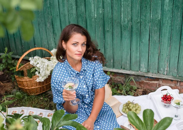 Summer picnic. A girl in a summer blue dress with polka dots and a straw hat sits on a picnic in nature and drinks mojito. Picnic with baguettes, grapes, cherries and cocktail.