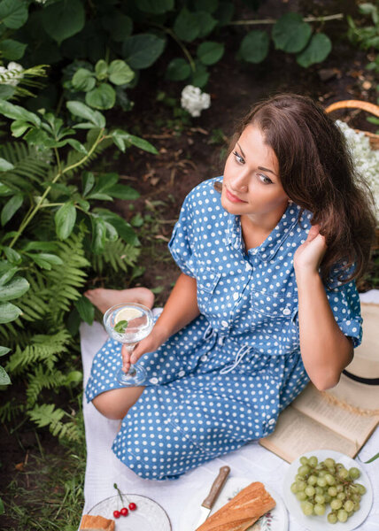 Summer picnic. A girl in a summer blue dress with polka dots and a straw hat sits on a picnic in nature and drinks mojito. Picnic with baguettes, grapes, cherries and cocktail.