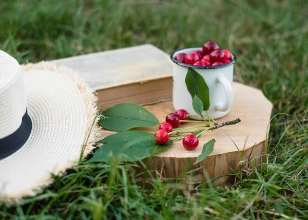 a cup with cherries, an old book, a summer straw hat on a background of green grass in the garden. Summer concept
