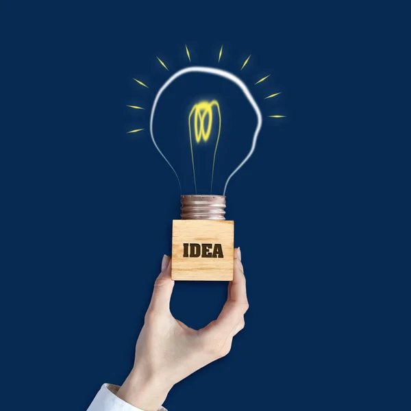 Idea concettuale. Mano femminile tiene un blocco di legno con l'iscrizione, Idea. Lampadina su sfondo blu. Il concetto di idee brillanti per le imprese, domande frequenti, ispirazione creativa — Foto Stock