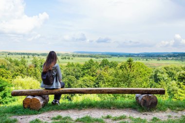 Sırt çantası olan kız, tepedeki bir bankta oturan güzel doğaya bakar. Doğaile yalnızlık.