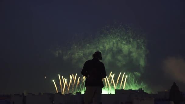 Sziluettjét fiatal srác kamerával tetőn állva. Tűzijátékok a háttérben — Stock videók