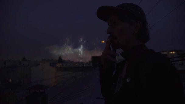 Jovem de pé no telhado do prédio fumar cigarro Fogos de artifício no fundo — Vídeo de Stock
