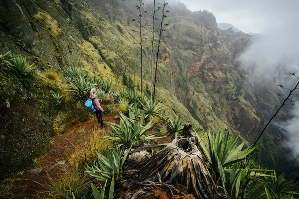 Женщина-путешественница, остановившаяся на краю вулкана над туманной зеленой долиной, заросшей агавами острова Санто-Антао в Кабо-Верде — стоковое фото