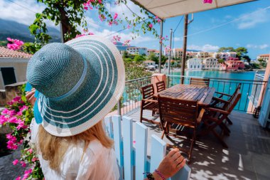 Assos village, Kefalonia, Yunanistan. Akdeniz'in turkuaz bay ve güzel renkli evler hayran rahat veranda teras kapısının önünde mavi sunhat kadın turist