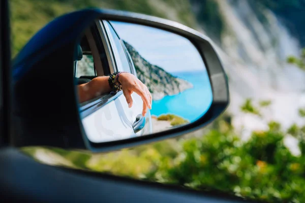 Gebräunte weibliche Hand im Seitenspiegel des Autos. blaues Mittelmeer und weiße Felsen Landschaft im Hintergrund — Stockfoto