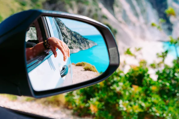 Braune weibliche Hand im Seitenspiegel des Autos. blaues Mittelmeer und weiße Felsen im Hintergrund — Stockfoto