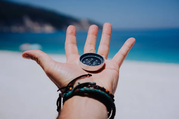 Outstretched hand holding black metal compass against white sandy beach and blue sea. Find your way or goal concept. Point of view pov