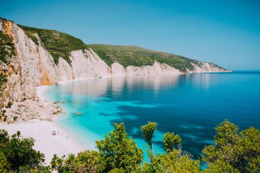 Sunny Fteri beach lagoon with rocky coastline, Kefalonia, Greece. Tourists under umbrella chill relax near clear blue emerald turquoise sea water clipart