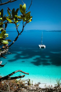 White catamaran yacht at anchor on clear azure surface with dark pattern in calm blue lagoon. Unrecognizable tourists relax on airbed in water near the beach clipart