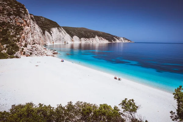 Playa de Fteri, Cefalonia, Grecia. Los turistas solitarios protegidos del frío sombrilla relajarse cerca de la laguna de agua de mar turquesa esmeralda azul claro. Enmarcado por el follaje del árbol — Foto de Stock