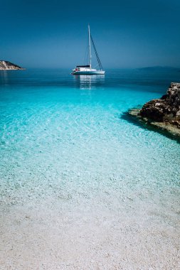 Yalnız beyaz Katamaran tekne drift sakin deniz yüzeyi yelken. Sığ su ve çakıl plajı ile saf gök temiz mavi lagün. Ön planda bazı kahverengi kaya taşları