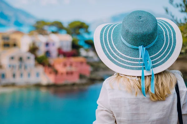 Visão traseira da mulher turista usar chapéu de sol azul e roupas brancas admirar vista de casas coloridas tranquilas coloridas da aldeia de Assos no dia ensolarado. Visitando Kefalonia no verão. Viagens de férias na Grécia — Fotografia de Stock