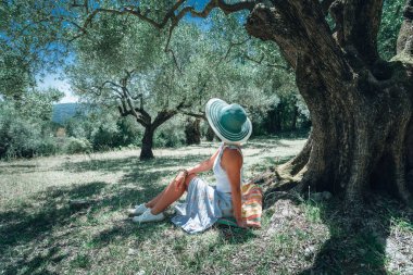 Genç kadın pastoral Akdeniz manzara arka plan üzerinde bir zeytin ağacının altında saman güneş şapkalı. Zeytin forest, peri masalı, rahatlatıcı, yaz, Yunanistan