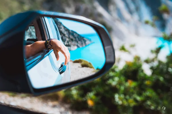 Female hand mirrored in the car side view mirror. Blue mediterranean sea and white rocks in background