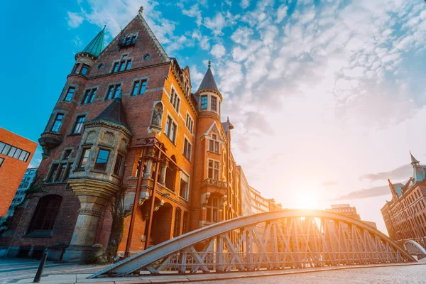 Ponte do arco do metal e edifício vermelho velho dos tijolos no distrito do armazém de Speicherstadt de Hamburgo HafenCity com luz do sunburst durante a hora dourada do por do sol e nuvens brancas de encontro ao céu azul acima — Fotografia de Stock